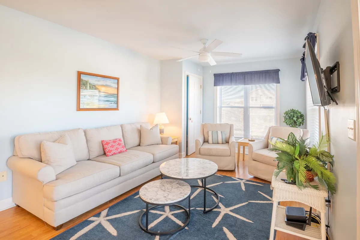 A second living room with a relaxed, welcoming ambiance. The room includes a comfortable sofa, armchairs, a coffee table, artwork on the wall, and soft window light, creating an inviting space for socializing or quiet moments.