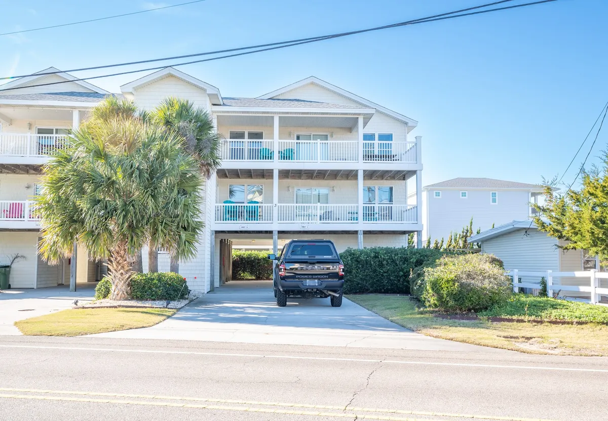 The image showcases the welcoming exterior of a spacious single-family house at Kure Beach, framed by a graceful palm tree and featuring a generous front porch. A black vehicle is parked in the drive, suggesting ample parking and easy access. The porch looks perfect for enjoying sunrise coffee or sunset views, and the palm tree adds a relaxed coastal vibe. The atmosphere is inviting, hinting at outdoor family gatherings and barbecues just steps from the sand.