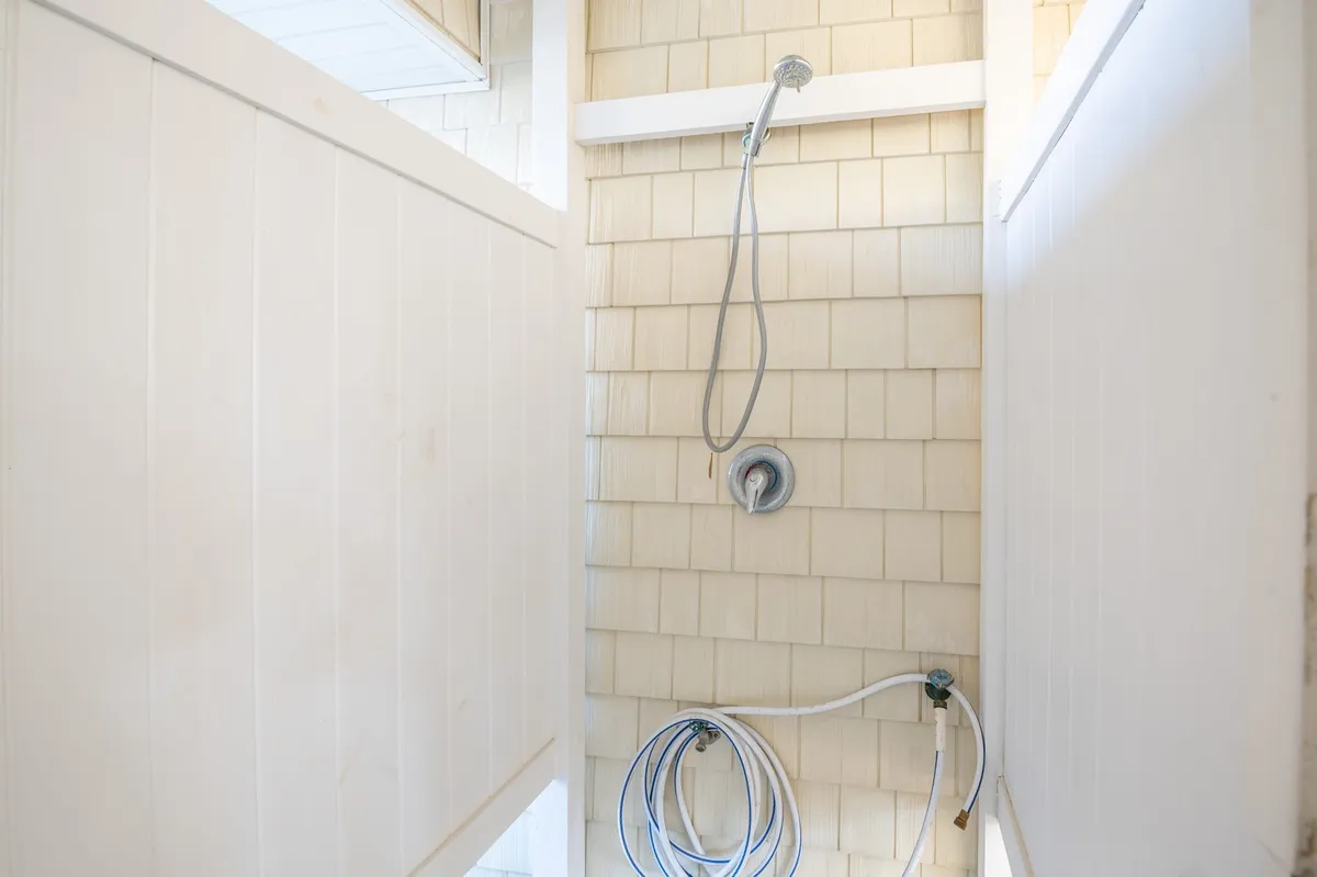 A private outdoor shower with a wooden shingle wall and white partitions offers a refreshing spot to rinse off after a day at the beach. The scene is bright and invigorating, with the functional and enjoyable outdoor amenity clearly on display.