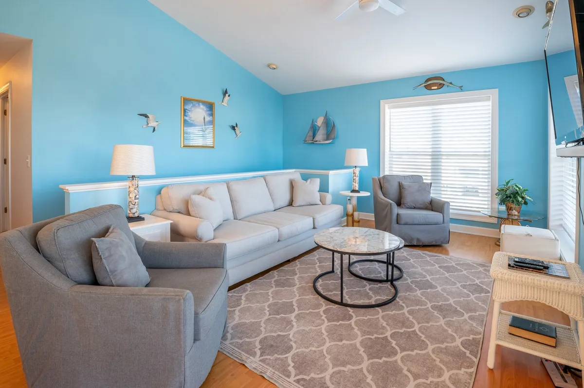A comfortable coastal-themed living space with a sofa, coffee table, wall art, and windows that let in natural light—ideal for socializing and enjoying sunrise or sunset views after outdoor activities.