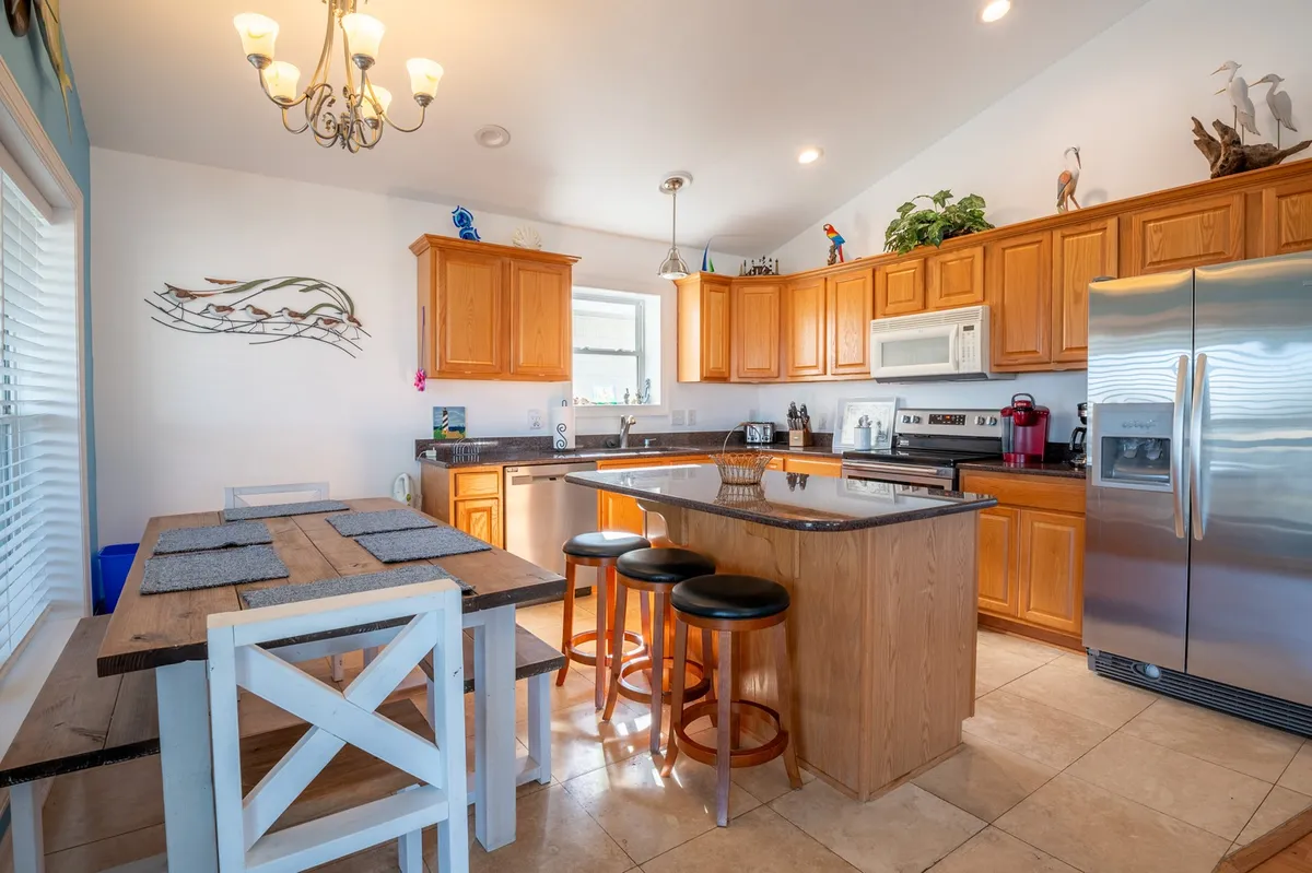 A spacious kitchen featuring a central island, stainless steel appliances, granite countertops, warm wooden cabinets, and decorative art. In the foreground is a dining table ready for a group meal, with ample lighting and an inviting atmosphere.