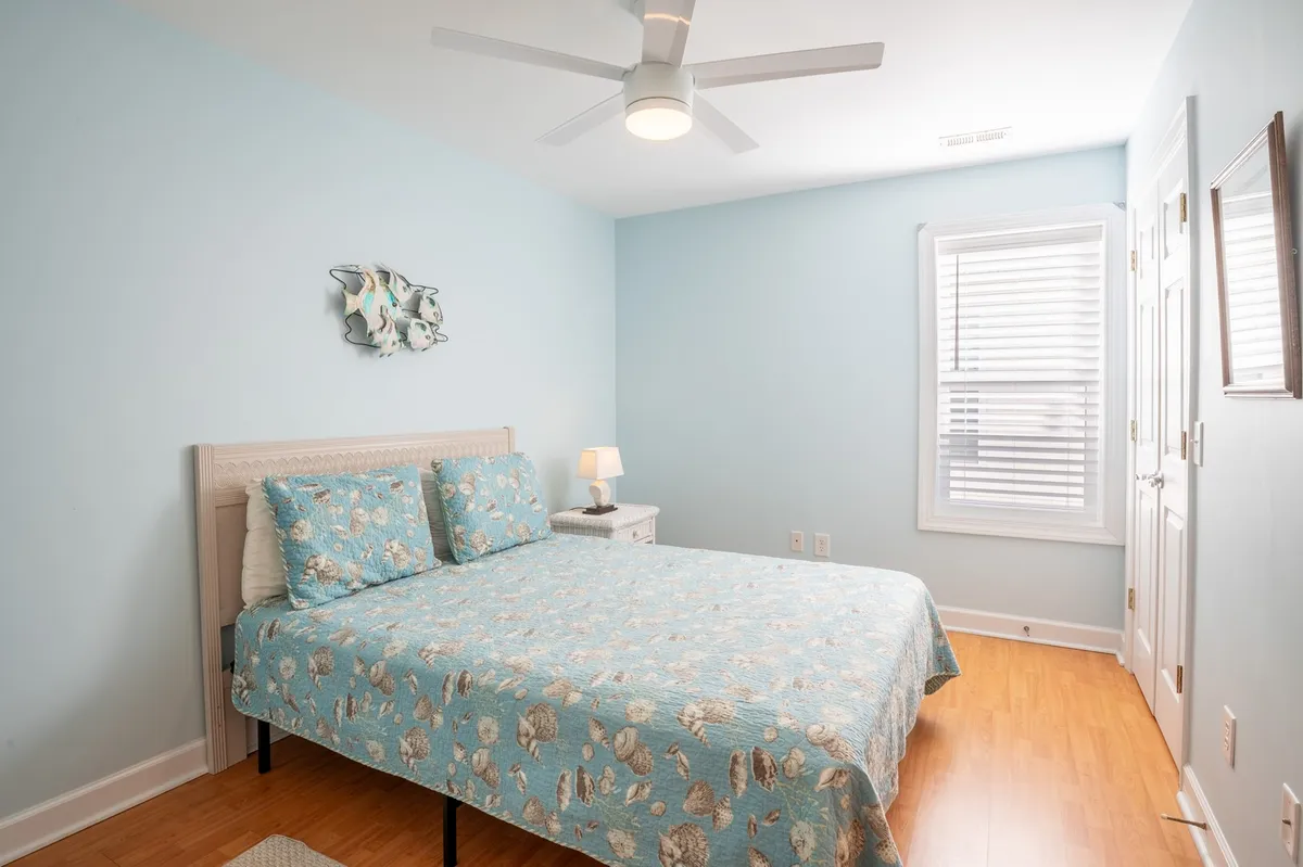 A tranquil bedroom featuring a bed with blue floral bedding, a nightstand with a lamp, and decorative wall art. Soft natural light streams in, creating a peaceful and inviting atmosphere, perfect for rest and relaxation.