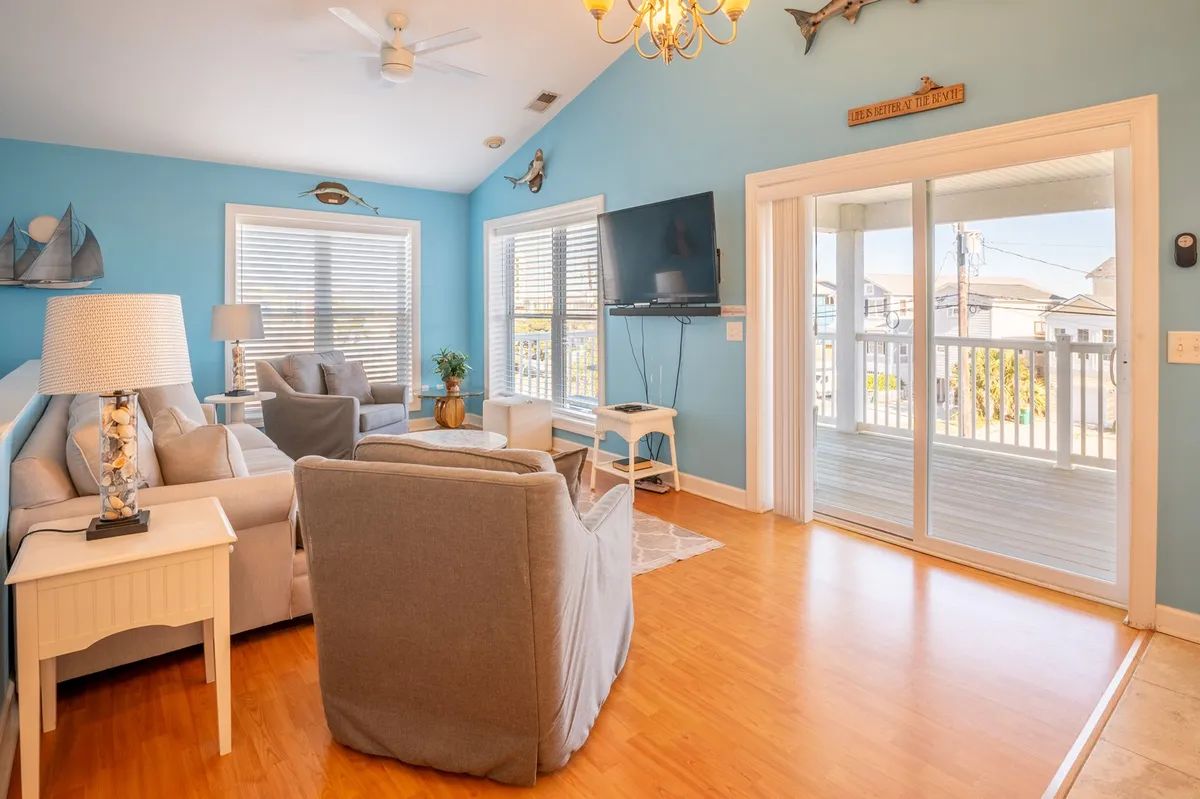 A bright, casual living room with large windows that offer glimpses of the outdoors. The setup includes comfortable sofa seating, a television, and ample space for socializing and relaxing after a day outside.