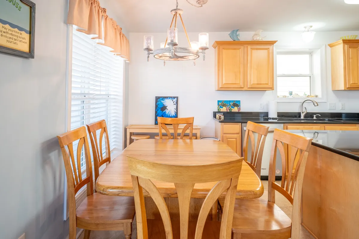 A warm dining area with a wooden table set under a chandelier, adjacent to a kitchen featuring cabinetry and tasteful wall art. The space is styled for shared meals and group gatherings, evoking comfort and community.