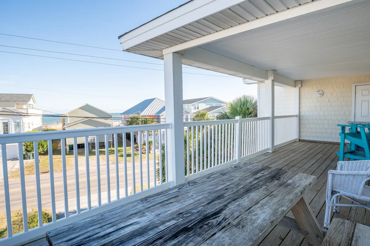 A spacious wooden deck with a sweeping view of the ocean, bordered by a white railing. The deck is furnished with comfortable outdoor seating, including Adirondack chairs and tables, creating an inviting spot to soak up the coastal atmosphere. Sunlight streams across the space, suggesting an ideal setting for both morning coffees and sunset gatherings.
