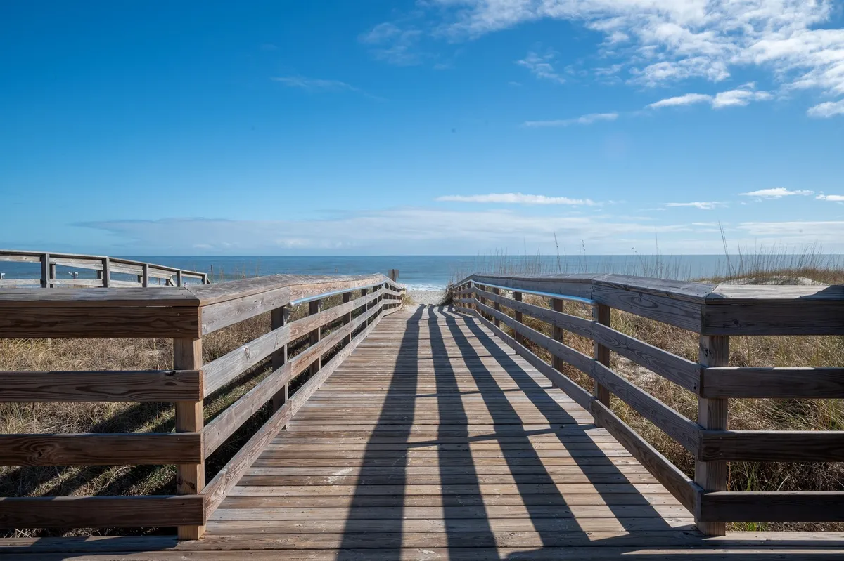 A wooden boardwalk stretches out towards the Atlantic Ocean, framed by a wide expanse of sand and a sky dotted with clouds. The scene captures the anticipation and ease of walking from the property directly to the beach, with unobstructed views of the water and natural beauty.