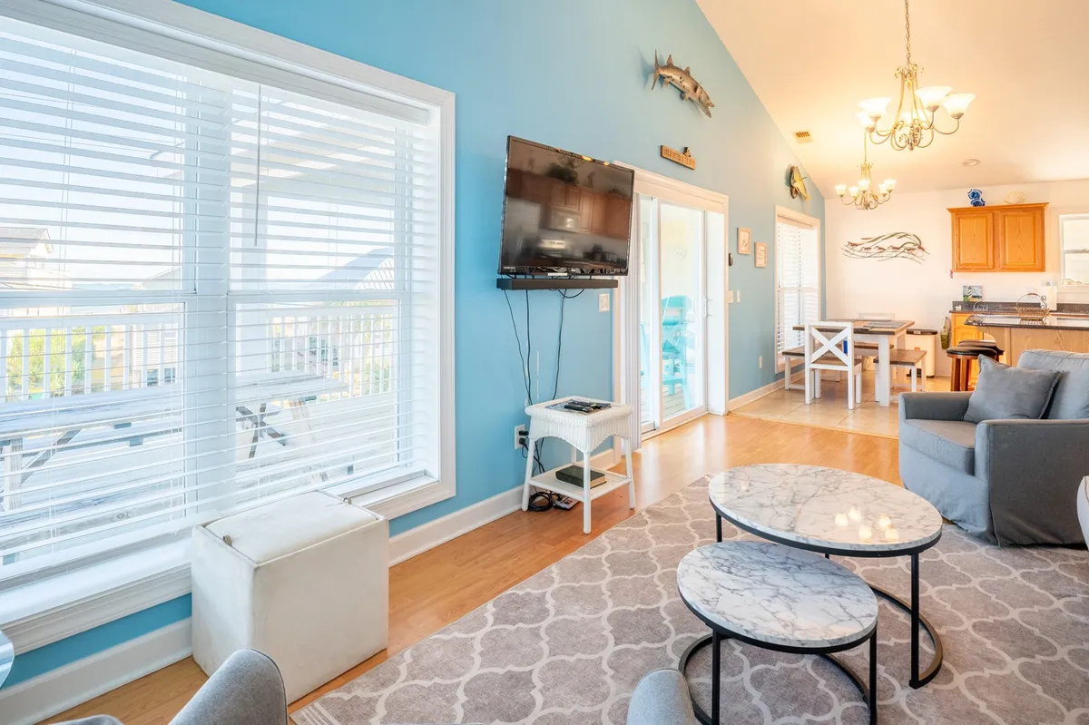 A bright and comfortable living room featuring large windows with blinds, a wall-mounted TV, a marble coffee table, and a cozy seating arrangement. The space is flooded with natural light, creating an inviting environment for family gatherings or relaxed afternoons.