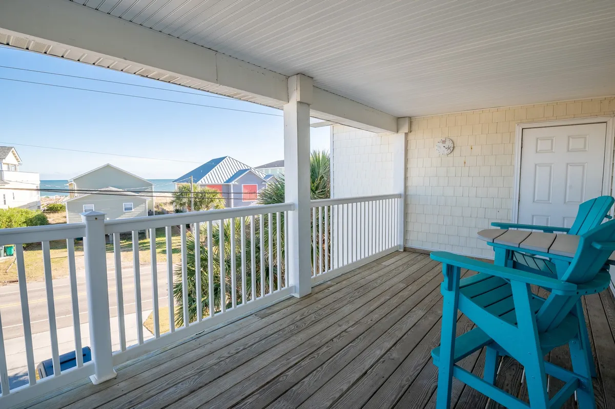 A wooden deck featuring several blue Adirondack chairs arranged to face the ocean, with a white railing framing the panoramic seascape. The tranquil setting invites quiet moments of relaxation, casual conversations, and an immersive experience of beach life.