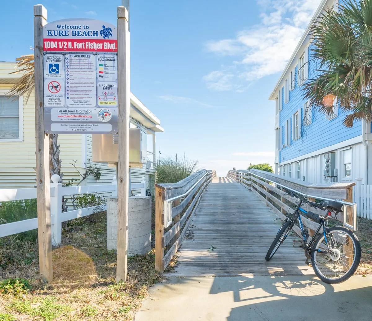 A welcoming beach access scene featuring a wooden walkway leading past a cheerful welcome sign, with a bicycle and lush greenery on either side. The view hints at easy, relaxed access to the coast and the outdoor activities nearby.